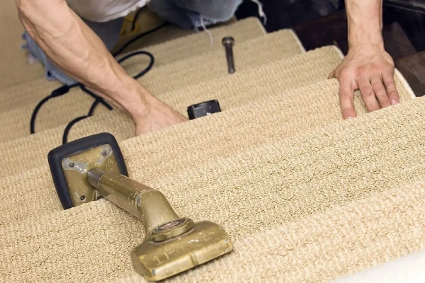 Professional performing sisal carpet installation on a staircase using a specialized knee kicker or carpet stretcher tool to ensure a tight fit on the tan-colored, looped carpet.